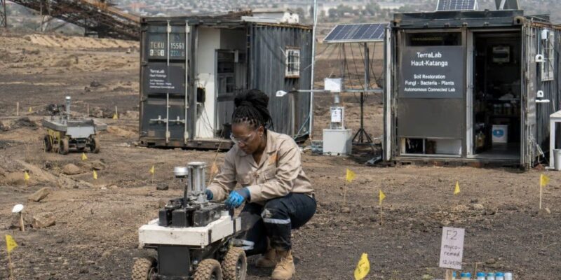 A woman next to a 4 wheel robot in a dusty field, next to containers with solar panels and lab equipment